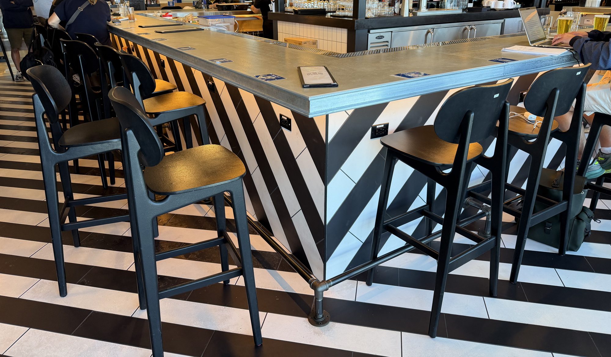 Corner of a zinc bar top lined with black bar stools at The Bar by Black Tap, JFK Terminal 8, New York
